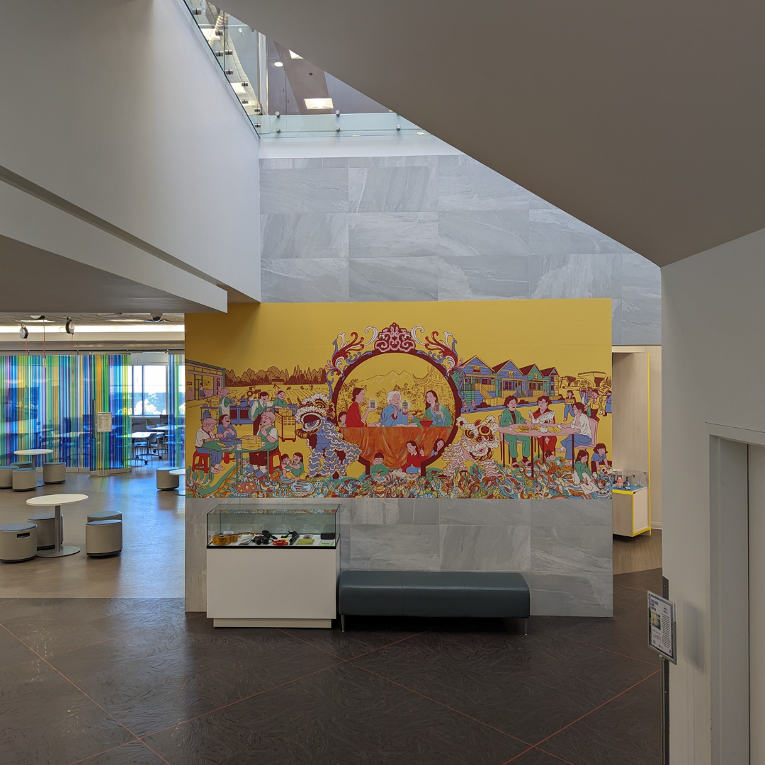 Installation view of the Suzhou Alley Women's Mural Exhibition at Brighouse Library. A cheerful and brightly coloured mural sits in the centre of the photo. In front is a waist-high glass display case. Next to it a gray upholstered bench.