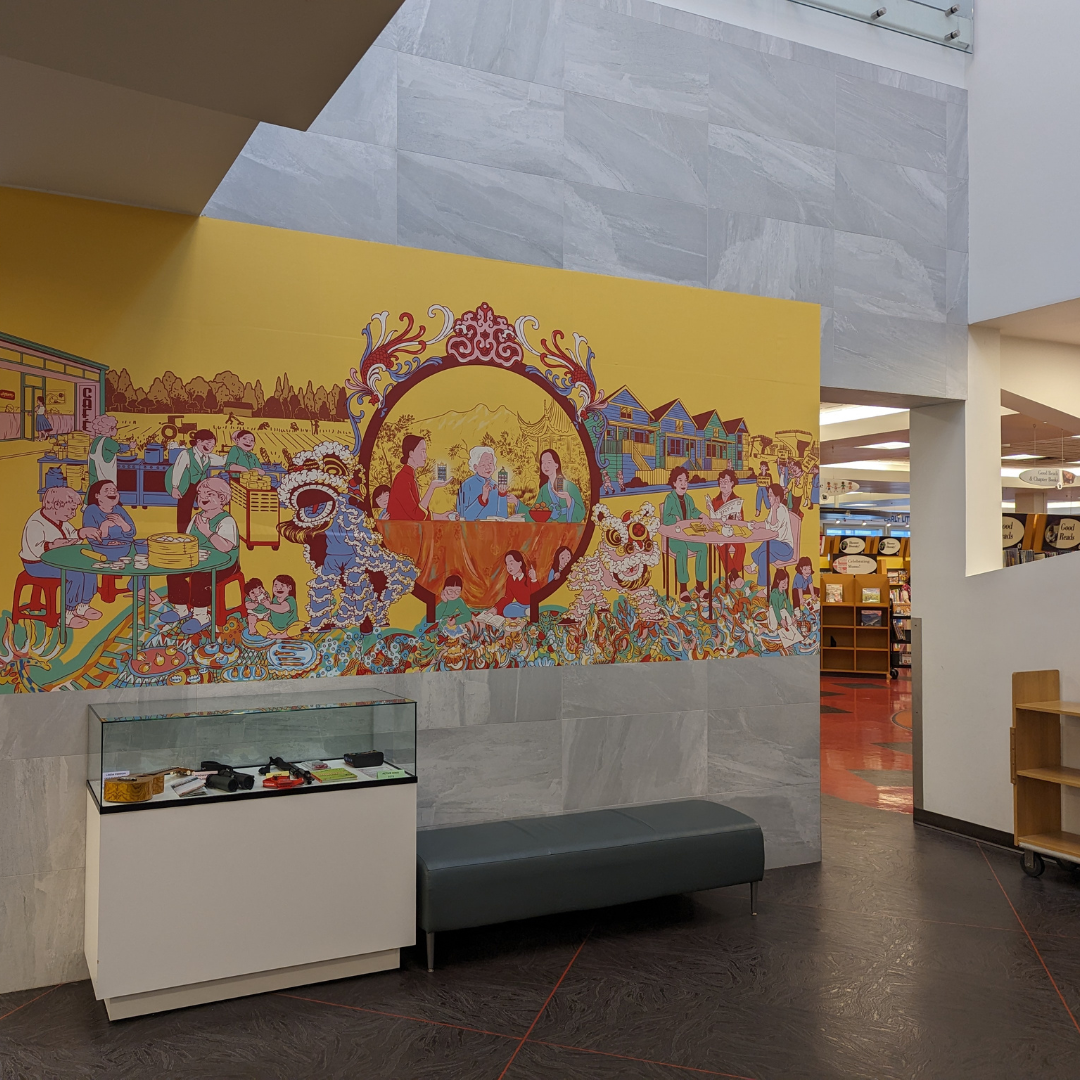 Installation view of the Suzhou Alley Women's Mural exhibition, side angle. On a yellow background, intricate drawings in cheerful colours shows scenes from the lives of women in Vancouver's Chinatown. The mural sits at eye level and fills the wall from left to right. To the left is an entryway leading to the library stacks.