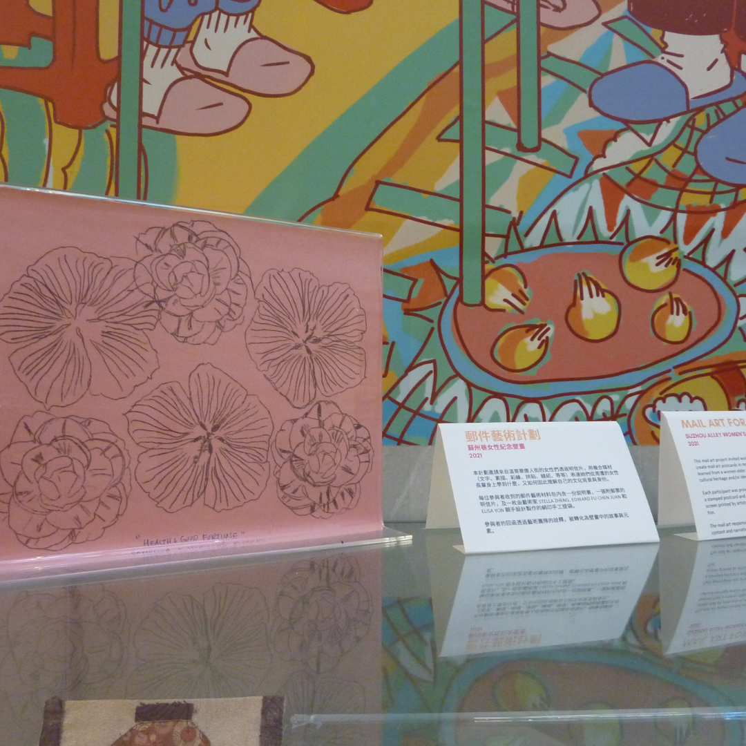 On top of a glass display case is a standing display of hand drawn flower stencils on a pink background. Next to it are two table cards describing the contents of the display case. One sign is in Chinese; the other in English. The mural is visible in the background.